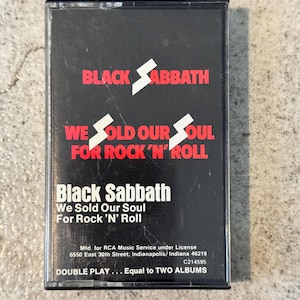 May include: A Black Sabbath cassette tape with a black case. The band's name and album title, "We Sold Our Soul For Rock 'N' Roll", are displayed in red and white. The text "DOUBLE PLAY... Equal to TWO ALBUMS" is also visible on the cassette.