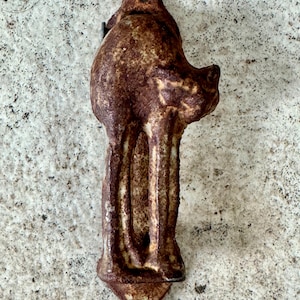 May include: A rustic, brown metal door pull shaped like a stylized animal. The pull has a weathered, textured appearance with visible rust and a small hole at the top and bottom for mounting. The design features a head and a long, vertical body.