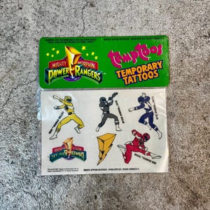 May include: A pack of Mighty Morphin Power Rangers temporary tattoos. The pack features a green background with the Power Rangers logo and the words "Temptoos Temporary Tattoos". The tattoos include images of the Power Rangers in their iconic costumes, including the Yellow Ranger, Black Ranger, Blue Ranger, and Red Ranger.