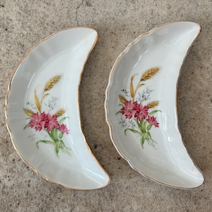 May include: Two white, crescent-shaped dishes with gold trim. Each dish features a floral design with pink flowers, green leaves, and wheat stalks. The dishes are likely for serving appetizers or small portions.