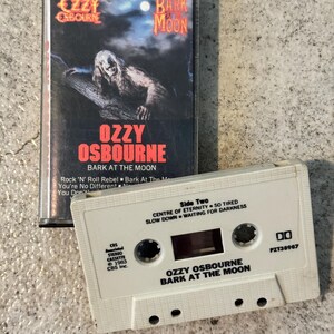 May include: A retro cassette tape of Ozzy Osbourne's "Bark at the Moon" album. The cover shows a werewolf figure under a night sky. The white cassette has song titles and album details printed on it, a classic music format.