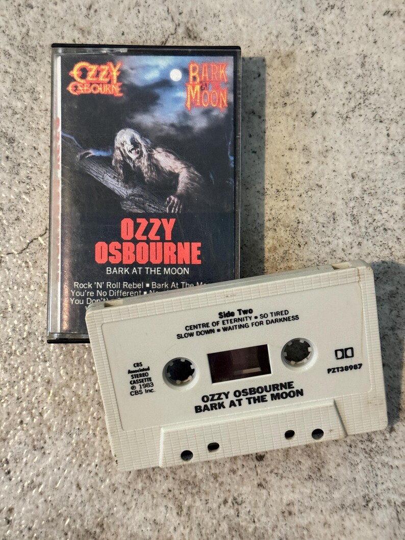 May include: A retro cassette tape of Ozzy Osbourne's "Bark at the Moon" album. The cover shows a werewolf figure under a night sky. The white cassette has song titles and album details printed on it, a classic music format.