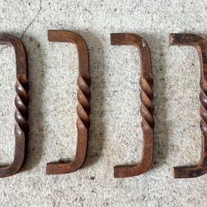 May include: Four rustic, L-shaped metal handles with a twisted, rope-like design on the vertical section. The handles are a weathered, reddish-brown color, suggesting an aged or antique appearance. They are arranged horizontally against a textured, light gray surface.