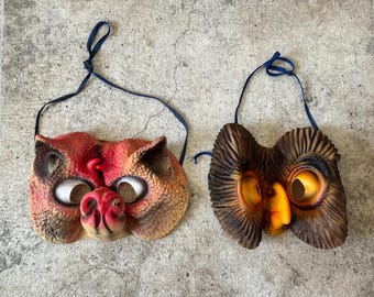 May include: Two vintage Halloween masks. One is a red and tan cat mask with white eyes and a red nose. The other is a brown and yellow owl mask with large yellow eyes and an orange beak. Both masks have blue string ties.