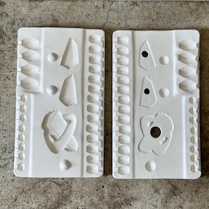 May include: Two white rectangular molds with various shaped cavities. One mold has oval, triangle, and abstract shapes. The other mold has circles, a flower shape, and other abstract shapes. The molds are on a textured, gray surface.