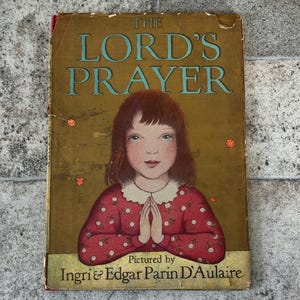 May include: Vintage book titled "The Lord's Prayer" with a gold cover and teal lettering. The cover features an illustration of a girl in a red dress with white polka dots, hands clasped in prayer. The book is pictured by Ingri & Edgar Parin D'Aulaire.