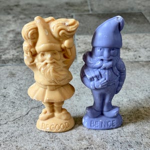 May include: Two small gnome figurines. One is tan and holds a bundle, the other is purple and has a satchel. The tan gnome's base says "BE GOOD", and the purple gnome's base says "BE NICE".