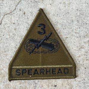 May include: Olive green triangular patch with black embroidered details. The patch features the number "3" above a tank and crossed cannons, with the word "SPEARHEAD" below. The patch is likely a military insignia.
