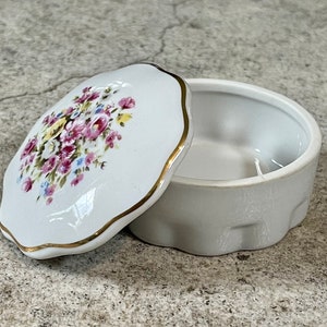 Op de afbeelding: Een witte porseleinen sieraden doos met een bloemenpatroon op de deksel. De deksel heeft een gegolfde rand en een gouden rand. De doos is leeg.