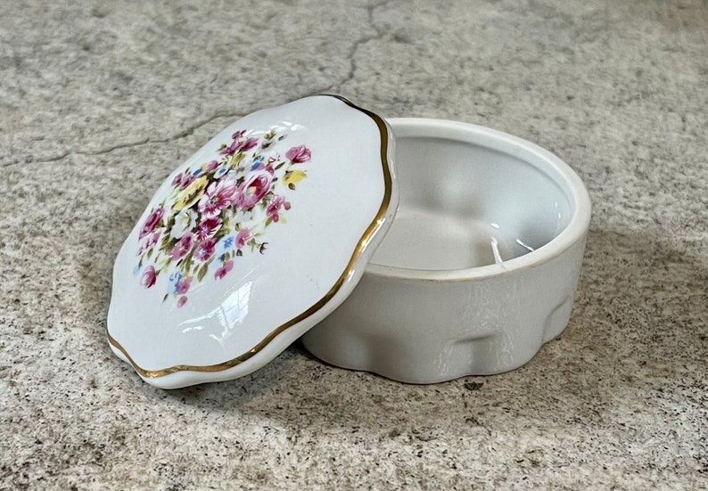 Op de afbeelding: Een witte porseleinen sieraden doos met een bloemenpatroon op de deksel. De deksel heeft een gegolfde rand en een gouden rand. De doos is leeg.