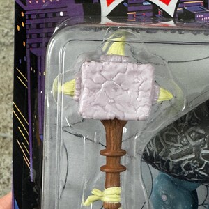 Puede incluir: Un martillo de juguete con una cabeza de color lila claro y un mango marr&oacute;n. El martillo forma parte de una figura de juguete, empaquetada en una funda de pl&aacute;stico transparente. El embalaje tiene un dise&ntilde;o de paisaje urbano y la palabra "SERIES!" en una explosi&oacute;n roja.