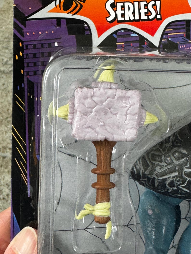 Puede incluir: Un martillo de juguete con una cabeza de color lila claro y un mango marr&oacute;n. El martillo forma parte de una figura de juguete, empaquetada en una funda de pl&aacute;stico transparente. El embalaje tiene un dise&ntilde;o de paisaje urbano y la palabra "SERIES!" en una explosi&oacute;n roja.