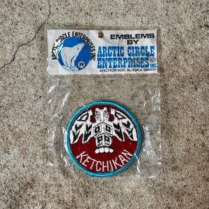 May include: A round patch with a red background and a teal border. The patch features a white and black design and the word "KETCHIKAN" in white. The patch is packaged in a clear plastic bag with the text "EMBLEMS BY ARCTIC CIRCLE ENTERPRISES INC."