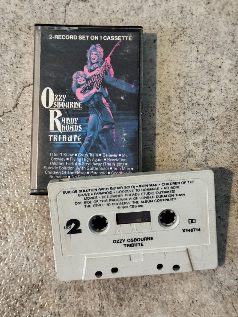 May include: A vintage cassette tape and case for the Ozzy Osbourne Randy Rhoads Tribute album. The case features a graphic of two musicians. The cassette tape is white with the album title and track listing.