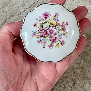 Op de afbeelding: Een wit porseleinen sieradendoosje met een gouden rand en een bloemmotief met roze, gele en blauwe bloemen.