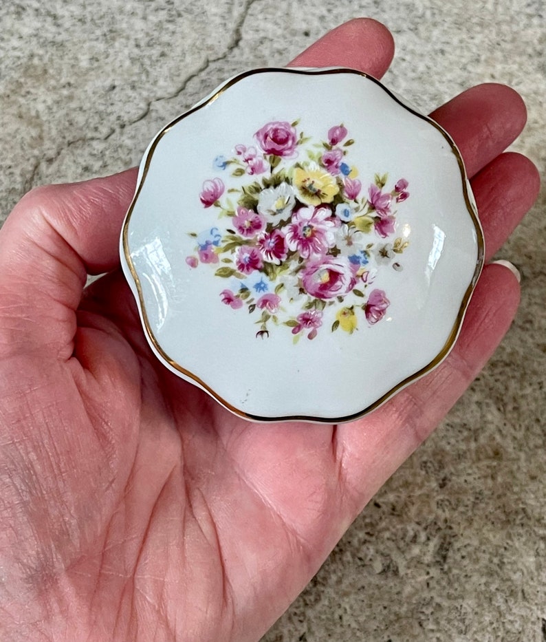 Op de afbeelding: Een wit porseleinen sieradendoosje met een gouden rand en een bloemmotief met roze, gele en blauwe bloemen.