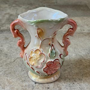 May include: A small, cream-colored ceramic vase with two ornate handles. The vase is decorated with embossed flowers in shades of yellow and pink, with green leaves. The handles and rim are accented with coral-colored paint.