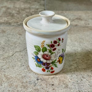 May include: A white ceramic jar with a lid, adorned with a floral design featuring red, blue, and yellow flowers. The jar has a cylindrical shape and a small handle on the lid. The overall design is vintage and decorative.