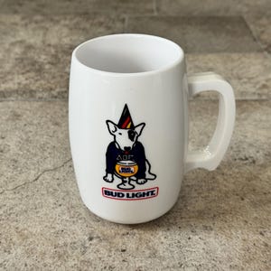 May include: White ceramic beer mug with a curved handle. The mug features a cartoon bull terrier wearing a party hat and a navy blue jacket, holding a glass of beer. The Bud Light logo is displayed at the bottom.