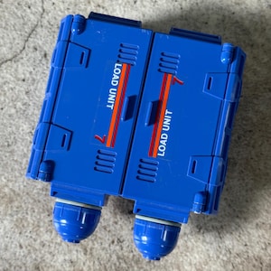 May include: A blue plastic toy with the words "LOAD UNIT" printed on it. The toy has a rectangular shape with two rounded sections at the bottom. Orange and red stripes accent the design.