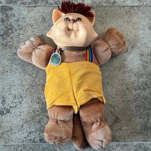 May include: A plush cat doll with a smiling face, brown fur, and a tuft of red hair. It wears yellow corduroy overalls with rainbow suspenders and a silver pendant. The doll's arms are outstretched.