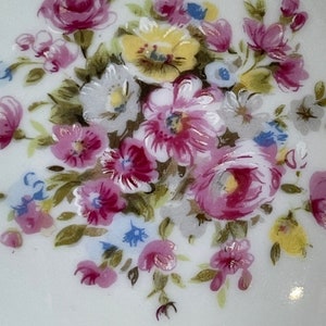 Op de afbeelding: Een wit keramiek bord met een bloemenontwerp. De bloemen zijn geschilderd in tinten roze, geel en blauw. Het ontwerp bevat een verscheidenheid aan bloemen, waaronder rozen, madeliefjes en vergeet-me-nietjes.