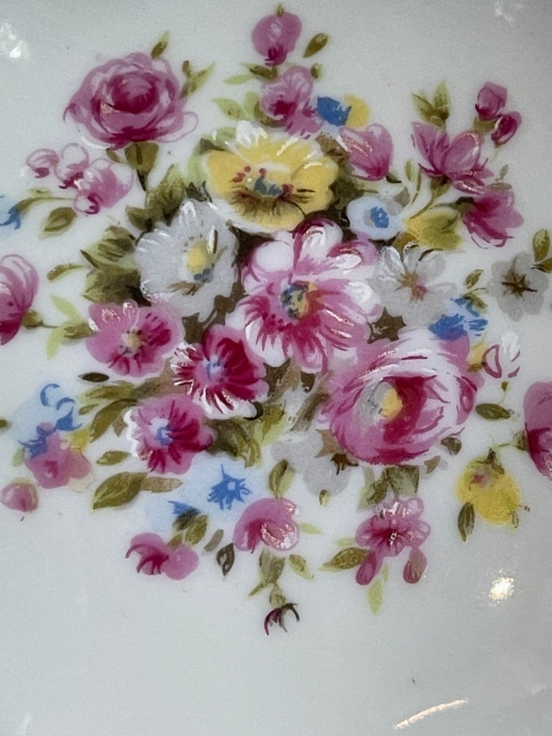 Op de afbeelding: Een wit keramiek bord met een bloemenontwerp. De bloemen zijn geschilderd in tinten roze, geel en blauw. Het ontwerp bevat een verscheidenheid aan bloemen, waaronder rozen, madeliefjes en vergeet-me-nietjes.