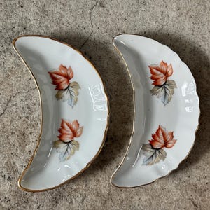 May include: Two white, crescent-shaped porcelain dishes with gold rims. Each dish features a painted design of a red and orange leaf with green and gray leaves. The dishes are on a gray surface.
