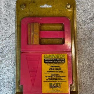 May include: A pink and yellow Nintendo game cartridge case with the text "ELIMINATOR NINTENDO SYSTEM SYSTEME NINTENDO" and "PREVENTS: POWER FLASHES! BLANK SCREENS! SCRAMBLED PICTURES! Assures Peak Performance From Your Nintendo System EMPÊCHE: LES FLASHES DE SURTENSION! LES ÉCRANS VIDES! LES IMAGES BROUILLÉES! Assure une performance optimale de votre système Nintendo."