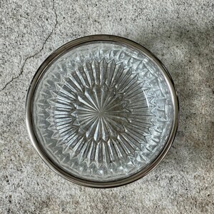 K&ouml;nnte beinhalten: Eine klare Glassch&uuml;ssel mit silbernem Rand und einem rautenf&ouml;rmigen Muster. Die Sch&uuml;ssel ist leer und steht auf einer grauen Betonfl&auml;che.