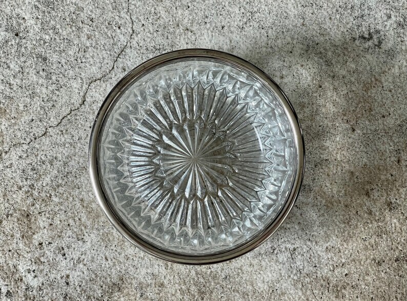 K&ouml;nnte beinhalten: Eine klare Glassch&uuml;ssel mit silbernem Rand und einem rautenf&ouml;rmigen Muster. Die Sch&uuml;ssel ist leer und steht auf einer grauen Betonfl&auml;che.