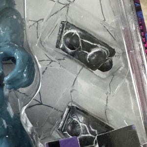 Puede incluir: Primer plano de un accesorio negro para figuras de acci&oacute;n, posiblemente un arma o herramienta, dentro de un embalaje de pl&aacute;stico transparente. El accesorio tiene una superficie texturizada detallada con formas redondeadas y una base rectangular. El embalaje tiene un fondo gris con un patr&oacute;n similar a una telara&ntilde;a.