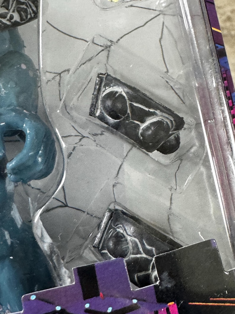 Puede incluir: Primer plano de un accesorio negro para figuras de acci&oacute;n, posiblemente un arma o herramienta, dentro de un embalaje de pl&aacute;stico transparente. El accesorio tiene una superficie texturizada detallada con formas redondeadas y una base rectangular. El embalaje tiene un fondo gris con un patr&oacute;n similar a una telara&ntilde;a.