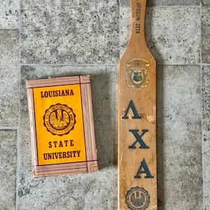 May include: A wooden paddle with the Greek letters Lambda, Chi, and Alpha, and a book with "Louisiana State University" printed on the cover. The paddle has a hole at the top and a crest design.