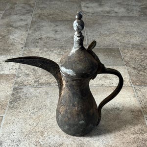 May include: A vintage, dark metal coffee pot with a long, curved spout and a handle. The pot has a bulbous body and a decorative top. The metal has a weathered appearance, with some areas showing lighter patches. The pot is likely used for serving coffee.