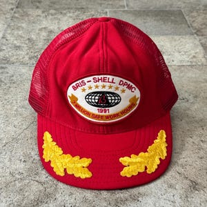 May include: Red trucker hat with a mesh back and a curved brim. The hat features a patch with the text "BRIS-SHELL DPMC" and "1991 Million Safe Work Hours". Gold leaf designs are embroidered on the brim.