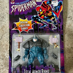 Puede incluir: Figura de acci&oacute;n Total Armor Rhino de la serie Special Collector, empaquetada en una tarjeta morada con gr&aacute;ficos de Spider-Man. La figura de Rhino es azul con armadura gris y accesorios. El embalaje incluye una advertencia sobre peligro de asfixia.