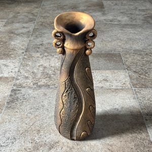 May include: A tall, decorative vase with a flared top and a textured, bronze-colored finish. The vase features intricate, swirling patterns and decorative elements near the top. The overall design is elegant and artistic, suitable for home decor.