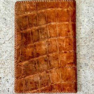 May include: A rectangular, brown leather item with a textured, crocodile-like pattern. The edges are stitched with a lighter brown thread. The item appears to be a wallet or small notebook cover.