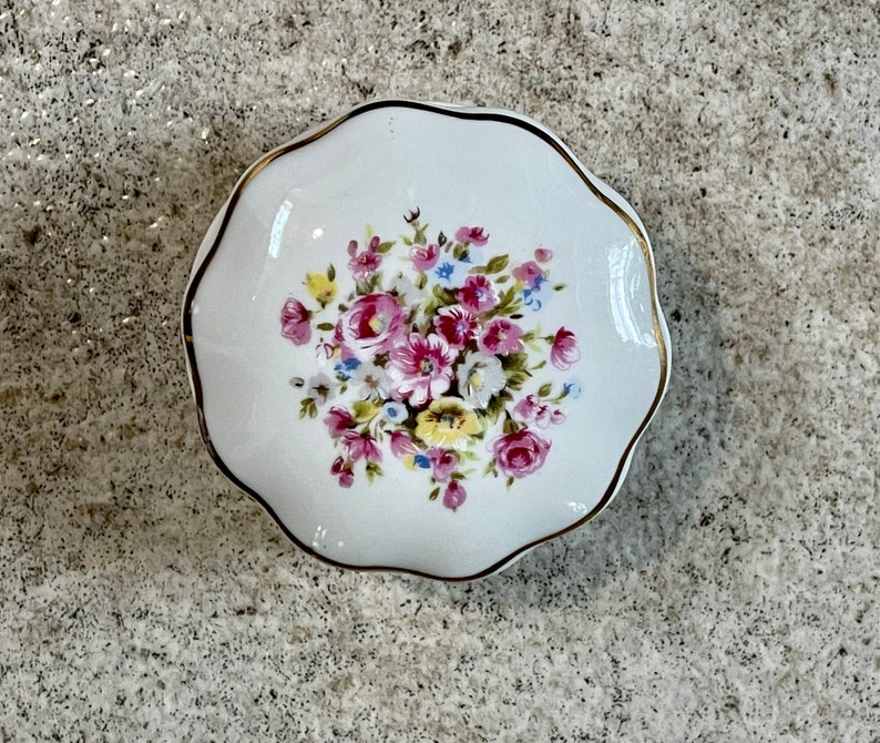 Op de afbeelding: Een wit porseleinen sieradenbakje met een gouden rand en een bloemmotief met roze, gele en blauwe bloemen.
