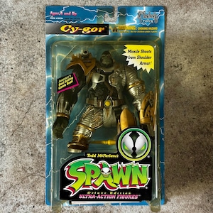 May include: A Spawn action figure of Cy-gor, a large, green, armored character with a missile launcher. The figure is packaged in a plastic blister pack with a blue and white background. The packaging features the Spawn logo and the text "Deluxe Edition Ultra-Action Figures".