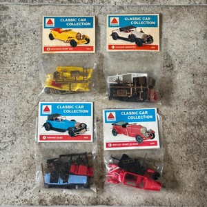 May include: Four vintage model car kits from the "Classic Car Collection" series. Each kit includes a car model and a card with the car's name and year. The cars are in various colors, including yellow, white, blue, and red.