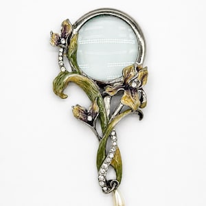 May include: A handheld magnifying glass pendant featuring a circular lens framed in dark metal. The handle and frame are decorated with enameled green and purple iris flowers, clear rhinestones, and a single dangling cream-colored faux pearl.
