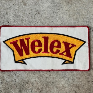May include: Embroidered patch with the word "Welex" in red letters on a yellow banner, outlined in black. The banner is set against a white background with a red border. The patch is rectangular.