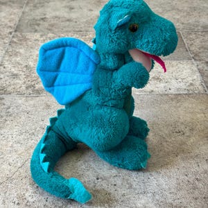 May include: A teal plush dragon toy with blue wings and a pink tongue. The dragon has a soft, textured body, small yellow eyes, and a long tail. The wings are a lighter shade of blue.