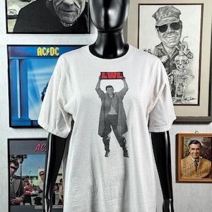 May include: A white short-sleeved t-shirt featuring a black and white graphic print of a person wearing a long coat and holding a sign above their head with the red text LWL. The shirt has a crew neckline and a standard fit on a black mannequin.