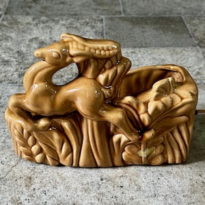 May include: A vintage ceramic planter in a tan color, featuring a deer leaping over a decorative base. The planter has a rectangular shape with detailed relief carvings of leaves and foliage. A power cord extends from the back.