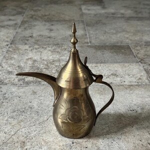 May include: A vintage brass coffee pot with a long spout, handle, and ornate floral design. The pot has a conical lid with a pointed finial. The surface has an aged patina, suggesting it is an antique or a well-loved item. The pot is likely used for serving coffee or tea.