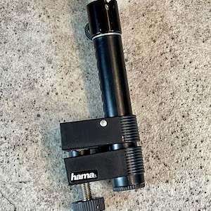 May include: Black camera mount with a clamp and a swivel head. The clamp has a screw to tighten it and the brand name "hama." is printed on the side.