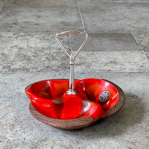 May include: A red and brown ceramic serving dish with a metal handle. The dish has three sections and is decorated with a swirl pattern. The dish is sitting on a gray tiled floor.
